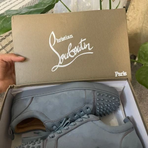Grå Christian Louboutin sneakers med nitar - Snygga grå sneakers från Christian Louboutin i mocka med ikoniska röda sulor och coola nitar framtill. Skorna har snörning och en stilren, modern look som sticker ut. Perfekta för dig som vill ha något unikt och trendigt i garderoben.