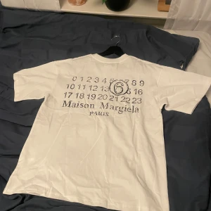 Maison Margiela vit t-shirt med tryck - Vit t-shirt från Maison Margiela med ikoniskt siffertryck och logga på ryggen. Framsidan har diskreta svarta stygn vid bröstet. Klassisk passform och rund halsringning. Tillverkad i mjuk bomull, perfekt för en clean och stilren look.