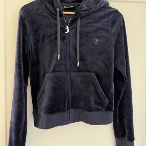 Mörkblå velour hoodie Juicy Couture - Säljer en mörkblå hoodie från Juicy Couture i mjuk velour. Tröjan har huva med snörning, dragkedja framtill och känguruficka. Perfekt för en chill och snygg look. Broderad JC-logga på bröstet och ribbade muddar.