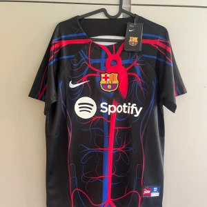FC Barcelona x Nike Patta fotbollströja - Unik FC Barcelona x Nike Patta fotbollströja i svart med färgstarka röda och blå ådror, klubbmärke på bröstet och Spotify-logga. Kortärmad modell i lätt, sportigt material. Perfekt för dig som vill sticka ut på planen eller läktaren.