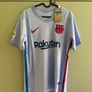 FC Barcelona bortatröja Nike M - Snygg FC Barcelona bortatröja från Nike i ljuslila polyester med klubbmärke och Nike-logga i rosa. Rak passform, kortärmad och med blå samt röda detaljer längs sidorna. Rakuten och UNICEF-loggor tryckta på framsidan och baksidan. Perfekt för dig som älskar Barca!