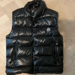 Svart dunväst från Moncler - Snygg svart dunväst från Moncler med glansig finish och klassisk logga på bröstet. Västen har hög krage, två dragkedjefickor och quiltad design. Insidan har en unik serietidningsdetalj. Perfekt för lager-på-lager och streetstyle.