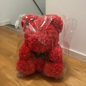 Rosbjörn - Bilderna visar en nallebjörn gjord av röda konstrosor, inslagen i plast. Det är ingen bok på bilderna, utan en dekorativ prydnad som passar perfekt som present eller inredningsdetalj.