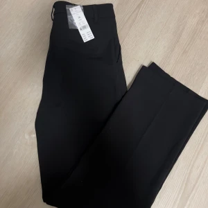 Svarta kostymbyxor från Gina Tricot med prislapp - Svarta kostymbyxor från Gina Tricot i rak modell. Byxorna har hällor för bälte, diskreta bakfickor och är tillverkade i en mix av polyester, viskos och elastan för en bekväm och stilren look. Perfekta för en clean och klassisk outfit.