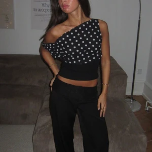 Design by Si prickig topp - Säljer en svart offshoulder topp med vita prickar och bred midjeresår. Toppen har en asymmetrisk, oneshoulder-design som ger en cool och trendig look. Perfekt att styla med vida byxor eller kjol för en snygg outfit.