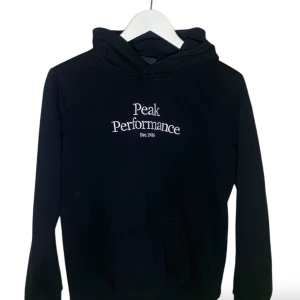 Svart hoodie från Peak Performance - Svart hoodie från Peak Performance med vit broderad logga på bröstet. Klassisk modell med huva och känguruficka framtill. Långärmad och i mjuk bomullsblandning, perfekt för en chill och sportig look.