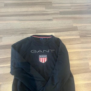 Svart GANT tröja med broderad logga - Svart långärmad tröja från GANT med broderad logga och sköld på bröstet. Klassisk rund halsringning med röd, vit och blå detalj på insidan av kragen. Perfekt för dig som gillar stilren och sportig look.
