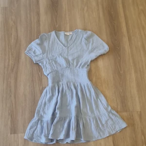 Ljusblå kortklänning från Gina Tricot - Supersöt ljusblå kortklänning från Gina Tricot med v-ringning, puffärmar och smock i midjan. Klänningen har volangkant nertill och är gjord i ett tunt, luftigt bomullstyg. Perfekt för varma dagar och ger en fräsch och somrig vibe.