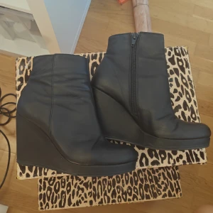 Svarta kilklackade stövletter i skinn - Snygga svarta stövletter från Bianco med kilklack och dragkedja på sidan. Skorna är tillverkade i skinn och har en stilren, minimalistisk design med rund tå och plattform. Perfekta för dig som vill ha höjd men ändå bekvämt.