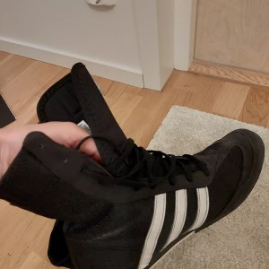Box hog 2.0 boots Adidas - Svarta höga sneakers från Adidas med klassiska vita ränder på sidorna. Skorna har snörning hela vägen upp och är tillverkade i ett lätt och ventilerande syntetmaterial. De är väldigt fina,  bekväma och användbara utan fel. Jag har bara använt ett par gånger (de är stora för mig).