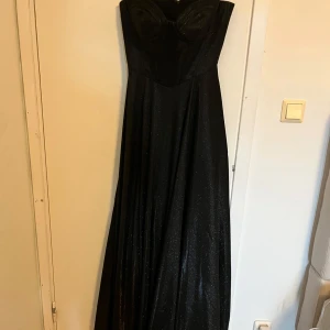 Svart glittrig balklänning från Dressroom linne - Supersnygg svart långklänning från Dressroom Linné med glittrigt tyg och markerad byst.  modell med figurnära överdel och vidd i kjolen. Perfekt för dig som vill sticka ut med en elegant look.