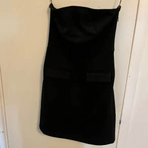 Svart axelbandslös klänning AWARE - Snygg svart axelbandslös kortklänning från AWARE. Klänningen har raka linjer, två dekorativa fickor framtill och en stilren look. Perfekt för dig som gillar minimalistisk stil och vill sticka ut med en klassisk svart klänning.