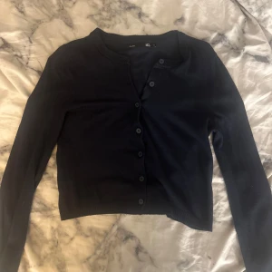 Svart kofta från 157, XS - En svart, klassisk kofta från lager 157 i storlek XS. Modellen är croppad med rund halsringning och knappar framtill. Långärmad och i mjukt, stickat material som passar perfekt nu till sommaren. Enkel och stilren design som funkar till allt.