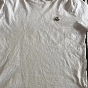 Vit Moncler t-shirt med logga - Snygg vit t-shirt från Moncler med klassisk rund hals och liten broderad logga på bröstet. Tillverkad i mjuk bomull som känns skön mot huden. Perfekt basic-plagg med exklusiv känsla och stilren design.