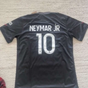 PSG Neymar Jr svart matchtröja - Snygg svart PSG-fotbollströja med Neymar Jr och nummer 10 på ryggen. Framsidan har Paris Saint-Germain-logga, Nike-märke och sponsortryck i vitt. Tröjan är kortärmad och gjord i ett lätt, sportigt material som andas. Perfekt för dig som älskar fotboll och Neymar.