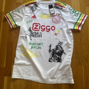Ajax x Bob Marley vit fotbollströja - Ajax x Bob Marley limited edition fotbollströja i vitt med färgglada ränder på ärmarna i rött, gult och grönt. Tryck med Ziggo-logga, Ajax-emblem och Bob Marley-grafik samt citat och illustrationer på både fram- och baksida. Tillverkad i lätt och ventilerande material.