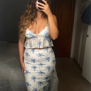 Vit långkjol och topp med blå blommor - Supersöt långkjol i vitt med blått blommönster. Kjolen är figurnära och har en romantisk vibe, perfekt för varma dagar. Materialet känns lätt och flowy, och mönstret är detaljerat med små blommor i olika blå nyanser. Jag har handsytt på fina pärlor och paljetter så därav blir varan dyrare då allt är handgjort. Ej använd.