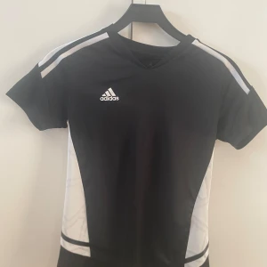 Svart och vit Adidas tränigströja XS - Snygg svart träningströja från Adidas i storlek XS. Tröjan har vita detaljer längs sidorna och axlarna samt Adidas-logga på bröstet. Tillverkad i ett lätt och ventilerande material som passar perfekt för träning.