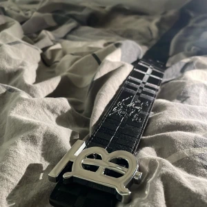 Burberry rutig skärp med silverdetalj - Snyggt Burberry-bälte med klassiskt rutigt mönster i beige, svart och röd. Skärpet har en stor, glänsande silverspänne med TB-logga och är gjort i präglat skinn. Perfekt accessoar för att lyfta din outfit med en lyxig touch. Och ej använt.