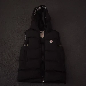 Svart dunväst med huva från Moncler - Säljer en svart dunväst från Moncler med stor huva och tydlig logga på bröstet. Västen har dragkedja framtill, två fickor med dragkedja och Moncler-tryck på insidan av huvan. Perfekt för lager-på-lager och streetstyle. Man kan skanna den.