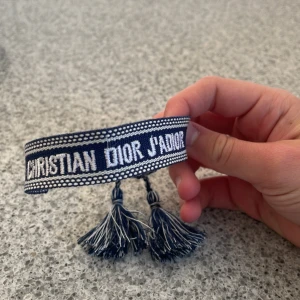 Blått Christian Dior J'Adior armband - Snyggt blått armband från Christian Dior med texten 'J'Adior' broderad i vitt. Armbandet har bred vävd design och avslutas med coola tofsar i blått och vitt. Perfekt statement piece för att lyfta din outfit.