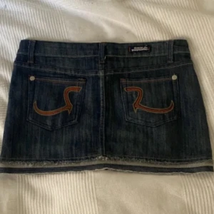 Jeanskjol från Rock & Republic - Mörkblå jeanskjol från Rock & Republic med rå kant nertill och klassiska fickor bak med snygg broderad detalj. Kjolen har femficksmodell, bälteshällor och silverfärgade nitar. Perfekt för en cool och avslappnad stil.