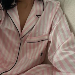 Ikonisk Victoria’s secret pyjamas - Snygg pyjamas från Victoria's Secret i satin material med rosa och vita ränder. Klassisk krage, svarta kontrastkanter och bröstficka med VS-logga. Perfekt för dig som gillar en söt och lyxig look. Aldrig sovit i men använd 1 gång! både byxor och skjorta, lånad bild men kan fixa egna