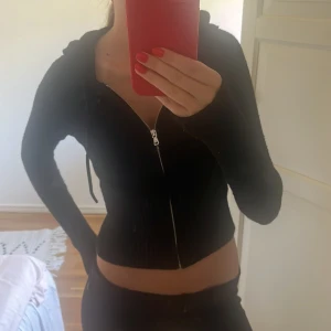 Svart croppad stickad hoodie - Svart croppad stickad hoodie med dragkedja framtill och huva. Jackan har lång ärm och ribbade detaljer vid midjan och ärmsluten. Perfekt för en chill och trendig look. Köpt för 500kr
