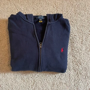 Mörkblå hoodie från Polo Ralph Lauren - Mörkblå hoodie från Polo Ralph Lauren med dragkedja och huva. Klassisk röd broderad logga på bröstet och mjukt material. Perfekt för chill dagar och enkel att matcha med jeans eller joggers.
