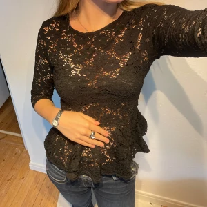 Svart spetsblus från Amisu - Svart blus från Amisu i genombruten spets med blommigt mönster. Modellen har trekvartsärm, markerad midja och lätt volang nertill. Dragkedja i ryggen för enkel påklädning. Perfekt för dig som gillar detaljer och en feminin look.