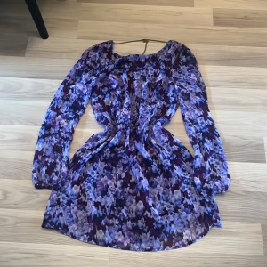 Lila blommig kortklänning med kedjedetalj - Supersöt kortklänning i lila och blå toner med blommigt mönster. Långärmad modell med markerad midja och snygg guldfärgad kedja i ryggen. Klänningen har ett glansigt innerfoder och flowigt tyg som ger en riktigt fin siluett.