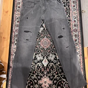 Grå Replay Anbass jeans med slitningar 34/32 - Säljer ett par grå Replay Anbass jeans med coola slitningar och trasiga detaljer framtill. Jeansen har klassisk femficksdesign, raka ben och snygga bakfickor med broderi. Perfekt för dig som gillar en avslappnad och trendig look.