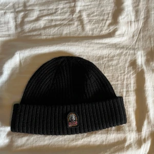 Svart ribbad mössa från Parajumpers - Svart ribbstickad mössa från Parajumpers med uppvikt kant och klassisk PJS-logga framtill. Snygg och enkel design som funkar till allt, tillverkad i mjukt material som håller dig varm under kalla dagar. Detta är den lite tjockare modellen av parajumper mössorna.
