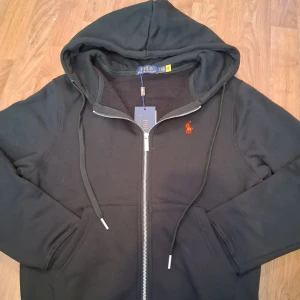 Svart hoodie från Polo Ralph Lauren - Svart hoodie från Polo Ralph Lauren med dragkedja och huva. Klassisk orange logga broderad på bröstet och snörning i huvan. Mjuk insida och praktiska fickor framtill. Perfekt för chill dagar eller när du vill ha en clean streetstyle.