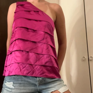 Rosa oneshoulder topp i satin - Säljer en snygg rosa oneshoulder topp med breda vågiga volanglager då den tyvärr är för stor för mig. Toppen har en glansig satinlook och är perfekt för dig som vill sticka ut. Den är ärmlös och har en asymmetrisk axel, vilket ger en cool och modern vibe. Perfekt till fest eller tjejkvällar!