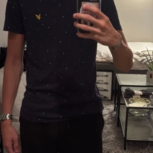 Lyle & scott - Snygg mörkblå t-shirt från Lyle & Scott med små vita och rosa/röda stjärnmönster över hela tröjan. Klassisk gul logga på bröstet och rund hals. Skön passform och tillverkad i mjuk bomull. Perfekt för dig som vill ha en stilren men ändå unik look. Storlek M, rätt slim fit