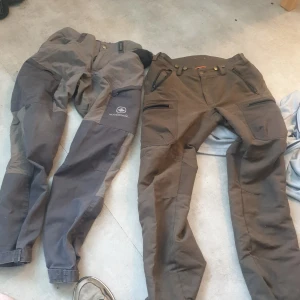 Två par outdoorbyxor Mackenzie & Kayoba - Säljer två par outdoorbyxor, ett par från Mackenzie och ett par från Kayoba Outdoor i storlek 48. Båda är i olika nyanser av grått och brunt, med flera dragkedjefickor och förstärkta partier på knän och bak. Perfekta för friluftsliv och äventyr.