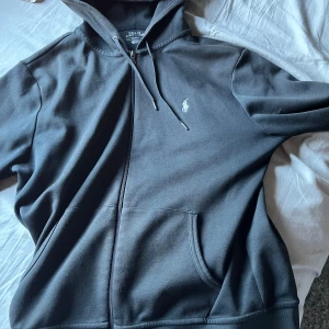 Svart zip hoodie från Polo Ralph Lauren - Svart zip hoodie från Polo Ralph Lauren med dragkedja och klassisk logga broderad på bröstet. Tröjan har huva med snörning och känguruficka framtill. Perfekt för en chill och stilren look.