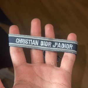 Christian Dior J'Adior armband - Snyggt vävt armband från Christian Dior med texten 'J'Adior' i vitt på mörkblå botten. Armbandet har justerbar knytning med tofsar i ändarna och coola kontrastdetaljer. Perfekt accessoar för att lyfta din outfit med en lyxig touch.