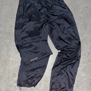 Svarta täckbyxor från Marmot - Svarta täckbyxor från Marmot i storlek L. Byxorna är tillverkade i 100% polyester och har elastisk midja samt dragkedjor längs benen. Perfekta för regniga eller blåsiga dagar när du vill hålla dig torr och bekväm.