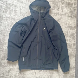 Svart Haglöfs Gore-Tex vindjacka - Svart vindjacka från Haglöfs i Gore-Tex-material med huva och hel dragkedja framtill. Jackan har två snedställda fickor med dragkedja och diskret logga på bröstet. Perfekt för blåsiga och regniga dagar, stilren och funktionell design.