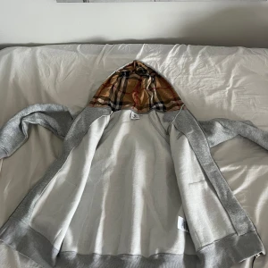 Grå zip-hoodie från Burberry - Grå hoodiejacka från Burberry med dragkedja och klassiskt rutigt foder i huvan. Jackan har kängurufickor framtill och är tillverkad i mjuk bomullsmix. Perfekt för dig som vill ha en stilren streetlook med en touch av lyx. Stolek M