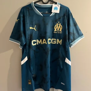 Marseille blå fotbollströja Puma - Snygg Marseille fotbollströja från Puma i mörkblå nyans med svarta detaljer och guldiga loggor. Kortärmad modell med vit krage och vita detaljer på ärmslut och sidor. Tillverkad i lätt och ventilerande material, perfekt för match eller träning.