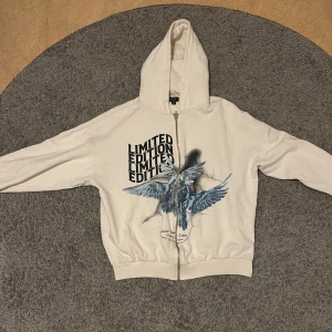 Vit hoodiejacka BoohooMAN M - Vit hoodiejacka från BoohooMAN i storlek M. Framsidan har tryck med texten 'LIMITED EDITION' och en blå fågelgrafik. Jackan har huva, hel dragkedja och ribbade muddar. Perfekt för dig som gillar streetstyle och unika prints.