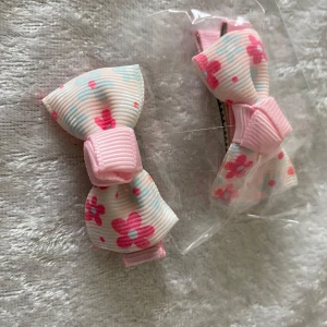 Söta hårrosetter med blommor & jordgubbar - Två par hårspännen med rosetter i pastellrosa nyanser. Ena paret har blommigt mönster i rosa och blått, andra paret har jordgubbsmotiv och prickar. Båda är gjorda i ribbad polyester och fästs med metallklämma. Perfekt för att piffa upp frisyren. Pris 25kr/par / bild , samma frakt 