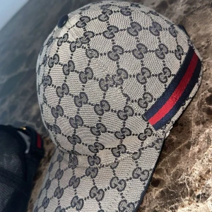 Gucci keps med GG-mönster och röd-blå rand - Säljer en exklusiv Gucci GG Supreme-keps i klassiskt mönster med den ikoniska röd-blåa randen på sidan. Kepsen är i mycket fint skick – sparsamt använd och välvårdad. Passar perfekt för dig som gillar stil, kvalitet och vill sticka ut med en tidlös accessoar. Kepsen är autentisk samt kvitto & box finns!	Äkthet: Garanterat äkta (kan visas vid köp) 	•	Nypris: Ca 3 800 kr. Pris kan diskuteras vid snabb affär!