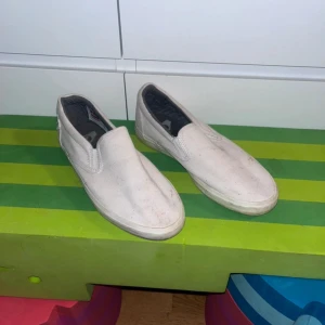 Vita slip-on sneakers från Quiksilver - Vita slip-on sneakers från Quiksilver i textil med gummisula. Skorna har en klassisk, enkel design med rund tå och platt sula. Perfekta för en avslappnad och stilren look. Lätta att ta på och av tack vare slip-on-modellen.