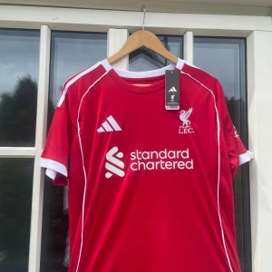 Liverpool Virgil #4 Adidas matchtröja - Liverpool FCs officiella röda matchtröja från Adidas med vita detaljer, klubbmärke och sponsortryck framtill. På ryggen står det 'VIRGIL' och nummer 4. Tröjan har korta ärmar och är tillverkad i lätt, ventilerande material.