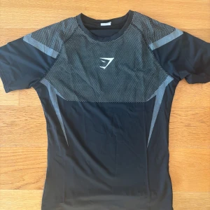 Svart Gymshark t-shirt med mönster - Säljer en svart och grå Gymshark t-shirt i funktionsmaterial, perfekt för träning. T-shirten har korta ärmar, sportig passform och coolt geometriskt mönster både fram och bak. Materialet är lätt och andas, med Gymshark-logga på bröstet.