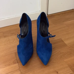 Blå mocka pumps från Zara - Snygga pumps i klarblå mocka från Zara med spetsig tå och hög klack. Skorna har en cool cut-out detalj på sidan och ett smalt spänne över foten. Perfekta för dig som vill sticka ut med färg och stil. Riktigt trendig siluett och elegant finish.